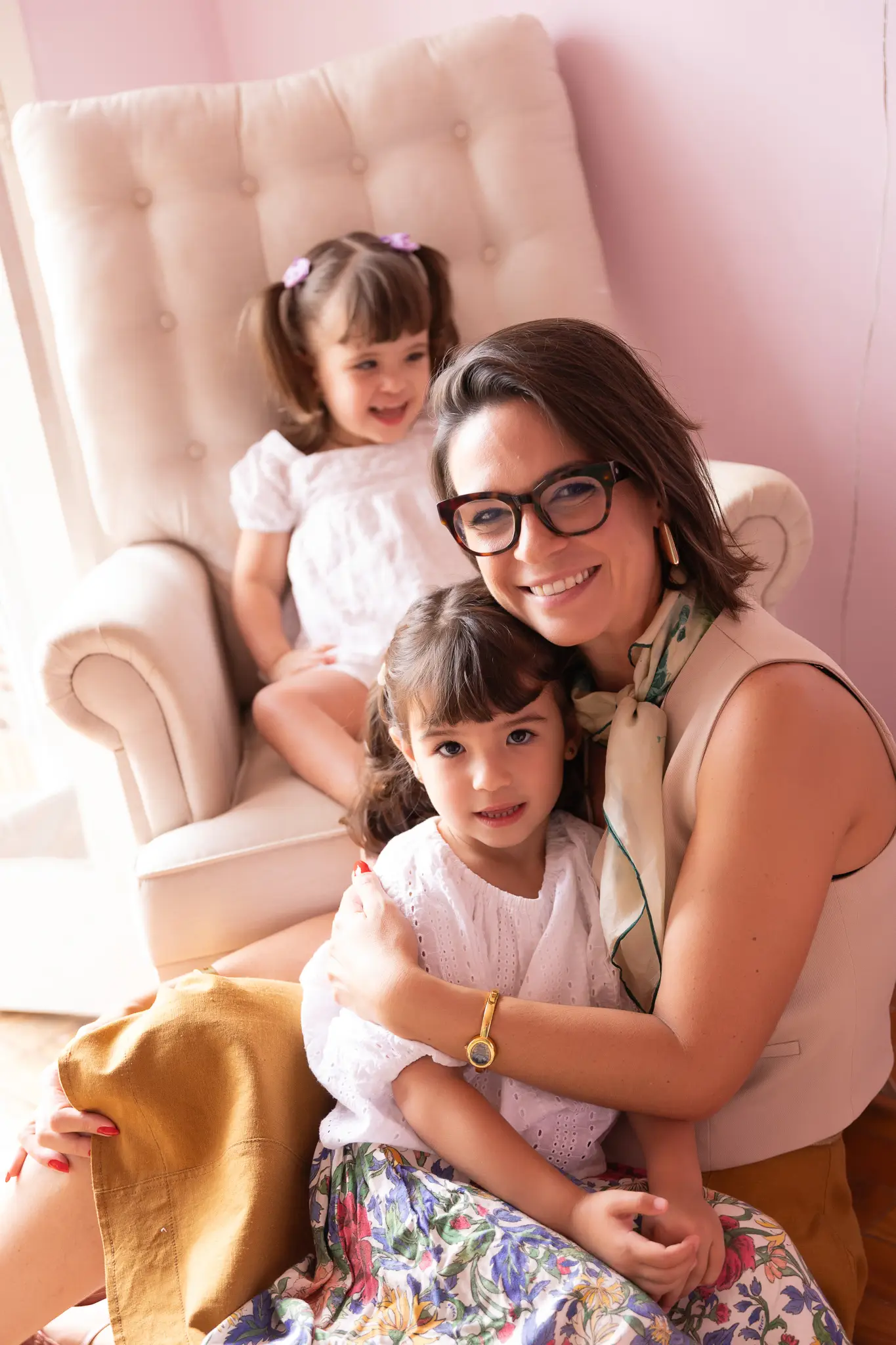 Nicole abraçada à filha mais velha, com a caçula sorrindo na poltrona atrás